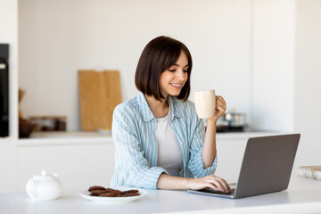 In the morning concept. Happy millennial woman using laptop and drinking hot coffee, sitting at table in kitchen