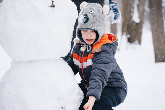 Children Play Outdoors In Snow. Outdoor Fun For Family Christmas Vacation. Two Little Kid Boy And Girl In Funny Hats Playing Outdoors. Happy Siblings Having Fun With Snowman