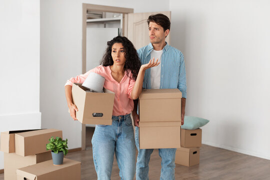 Confused couple holding cardboard boxes, walking in new house