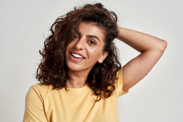 Pretty young woman with dark curly hair in casual wear looking cheerful at camera isolated over gray background