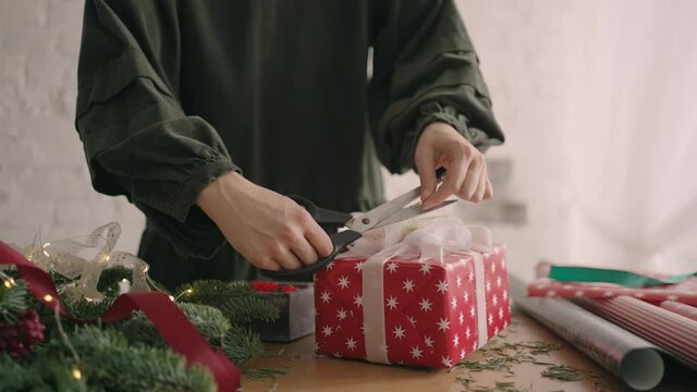 Close-up: Crop hands decorating wrapped box with string. Female packing cardboard gift box on wooden table with various decorative items prepared for Christmas celebration.