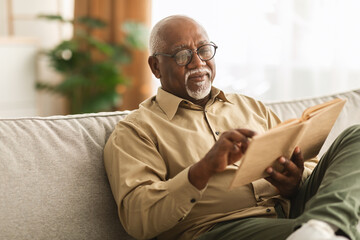 Senior African Man Reading Book Sitting On Couch At Home - obrazy, fototapety, plakaty