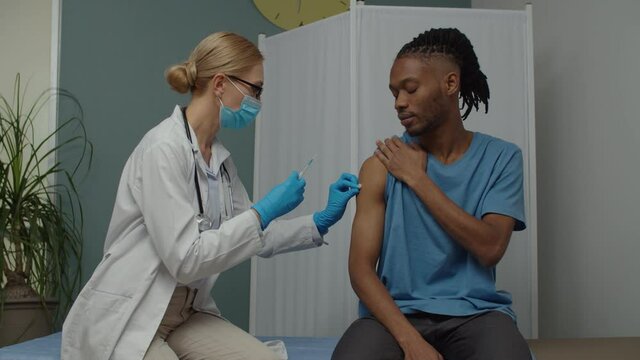 Attractive young adult man taking vaccination shot, sitting on diagnostic bed indoors. Female doctor injecting medicine or vaccine with needle to male patient in medical center
