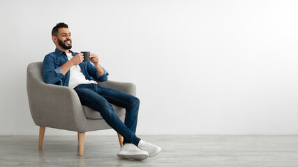 Full length of young Arab man drinking hot coffee in armchair against white studio wall, banner with free space