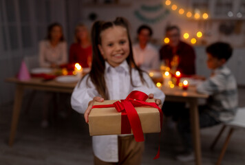 Adorable little girl giving wrapped gift box at camera during festive dinner at home, selective focus
