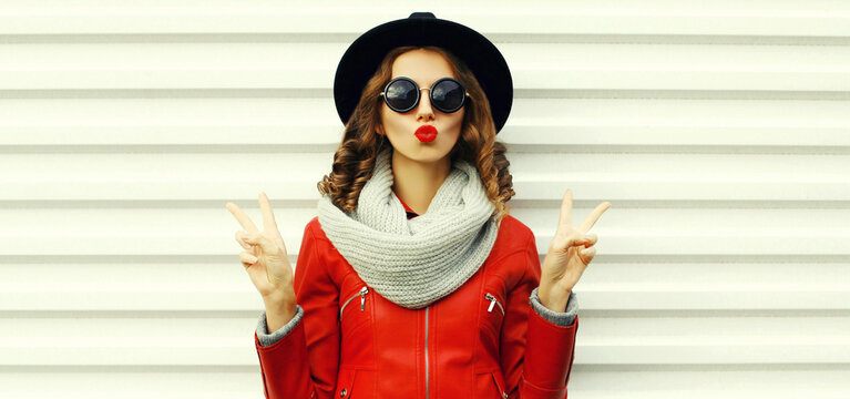 Portrait of beautiful young woman blowing her lips sending sweet air kiss posing wearing a red jacket, black round hat on white background