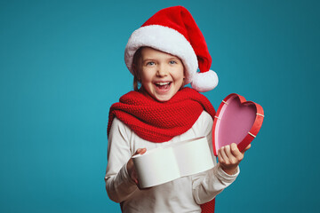 Happy little girl wearing christmas hat and red scarf opening a gift box isolated on blue background. Surprised cute kid with a wrapped present