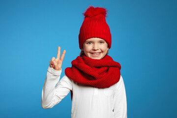 Cheerful cute child wearing red hat and scarf, looking at camera and showing peace gesture while standing isolated over blue background 