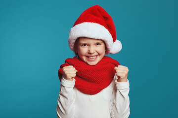 Cheerful little girl wearing christmas hat and red scarf raising fists up and celebrating over blue background 