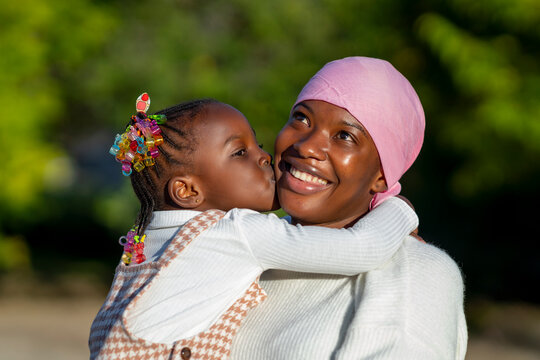 Cute African kid kissing cheek of smiling mother against green trees