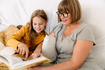 White girl reading book with her grandmother while sitting on sofa