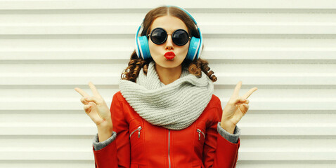 Portrait of stylish modern young woman listening to music in headphones blowing her lips wearing a red jacket on white background