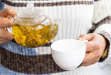 granny,senior woman in striped sweater pullover holds pouring herbal green plant tea from a transparent glass kettle.granny and nephew hands palms holding cup with herbal tea. white ceramic cup. cozy 