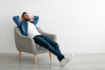 Calm young Arab man relaxing in armchair against white studio wall, copy space