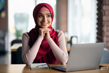 Excited young woman in hijab working on laptop, cafe interior