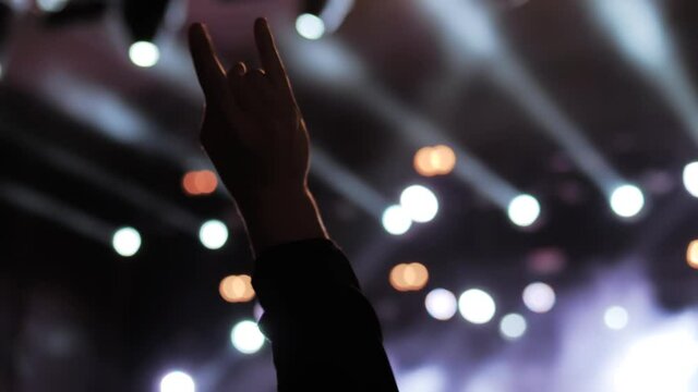 Close up: man partying, raising hands up, showing sign - devil horns gesture at rock concert in front of stage of nightclub. Colorful stage lighting. Nightlife and entertainment concept