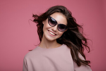 High fashion photo of a beautiful elegant young woman in a pretty pink jumpsuit, sunglasses posing on pink background. Slim figure. Studio Shot. Luxurious hair, shiny curls. Femininity and tenderness