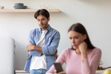 Obraz premium Displeasure offended sad young european husband looks at his busy wife, chatting in smartphone after quarrel