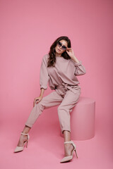 High fashion photo of beautiful elegant young woman in a pretty pink jumpsuit, sunglasses posing on studio background. Slim figure. Model sits on cube cylinder. Monochrome. Luxurious hair, shiny curls