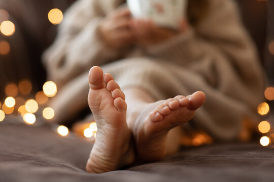 Children bare feet and hands close up garland lights. flaffy fuzzy warm knitted beige sweater. Christmas concept, holiday.Happy New Year.Selective focus. Child girl sitting on sofa inside.Cozy vibes