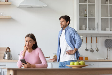 Sad young caucasian wife surfing in internet in smartphone, ignoring upset offended husband