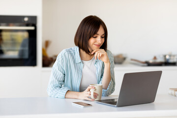 Remote job oppurtunities. Excited lady working on laptop and drinking morning coffee, sitting in kitchen, free space