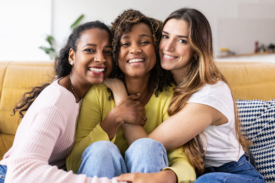 Three united beautiful smiling women sitting together on couch - Portrait of young multiracial girl friends hugging each other and relaxing at home - Friendship, youth and millennial people concept
