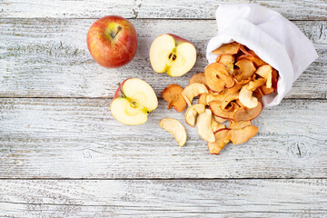 A pile of dried slices of apples pills out of a white pouch and fresh ripe apples on wooden background.