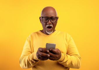 Emotional senior African American man in glasses looking at his smartphone in shock on orange studio background