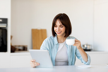 Positive caucasian woman drinking coffee and using digital tablet, surfing internet online, sitting at table in kitchen