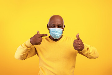 Senior black man pointing at mask, showing thumb up, recommending facial protection during...