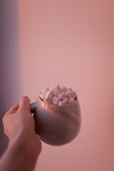 Hand holding a cup with cocoa with marshmallows with pink sunset light. Minimalist shot of winter drink.