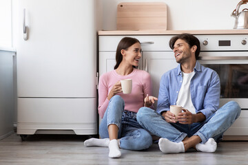 Smiling young european lady and guy with cups drink coffee in morning sitting on floor,...