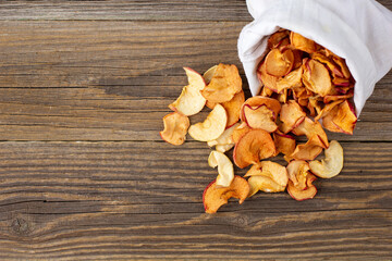 A pile of dried slices of apples pills out of a white pouch on wooden background. Dried fruit chips. Healthy food