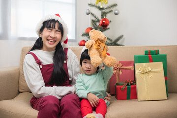 Obraz na płótnie Canvas Asian family Mother giving the teddy bear to child. Happy moment Mom and son in christmas festival. They dressing christmas theme with decorative pile gift box on sofa. Giving present each other.