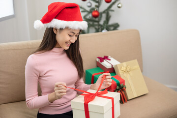 Beautiful Asian woman unpack the present gift box pull ribbon guess what inside the box. Young Happy cute girl celebrate in christmas holiday festival with many gift box sitting on sofa at home