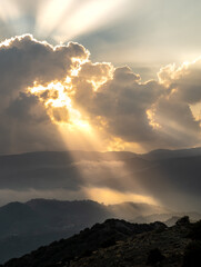 Bright sun rays light shining through dark clouds over mountain at sunset. Dramatic sky in winter