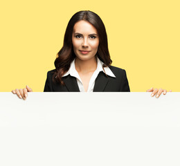 Naklejka premium Happy smiling brunette business woman in confident black suit, standing behind, peeping from blank banner or mock up signboard, isolated over yellow color background.