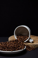 Cup filled with coffee beans on a black background. Freshly roasted coffee beans are poured out of the cup. Excessive consumption of caffeine