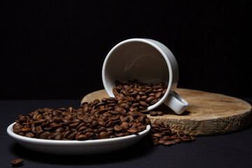 Cup filled with coffee beans on a black background. Freshly roasted coffee beans are poured out of the cup. Excessive consumption of caffeine