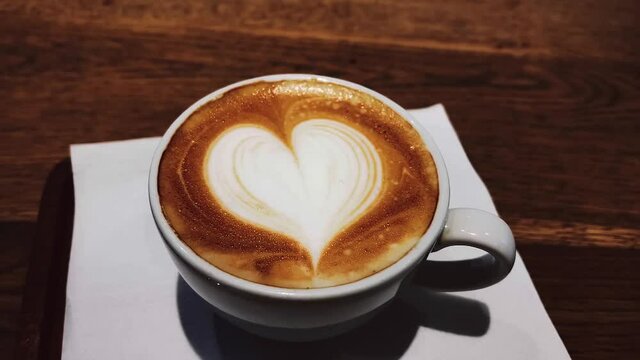 Coffee break and romantic mood concept. Cup of caramel cappuccino with heart shaped foam art made of lactose-free milk, served on wooden table in cafeteria. High quality 4k footage