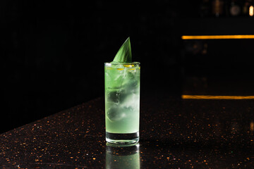 A horizontal photo of a light green alcoholic cocktail in a highball glass with ice, garnished with...