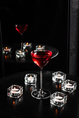 A red alcoholic cocktail in a nick and nora glass with a reflection in the mirror, candles, black table