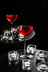 A red alcoholic cocktail in a nick and nora glass with a reflection in the mirror, candles, black table