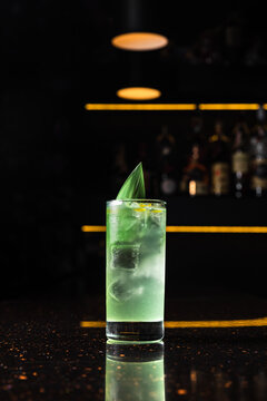 A light green alcoholic cocktail in a highball glass with ice, garnished with a bamboo leaf, at the bar counter, dark background