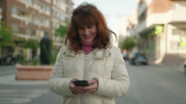 Middle age redhead woman smiling confident using smartphone at street