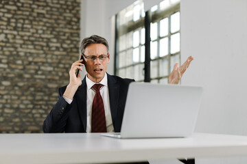 Frustrated businessman communicating with someone over cell phone while working on a computer