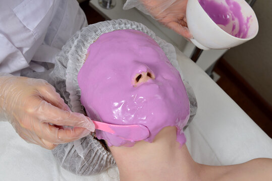 Cosmetologist applying alginate mask on young woman's face in beauty salon. Alginate mask for skin. Cosmetologist applying alginate mask on young woman's face in beauty salon. Lilac Mask