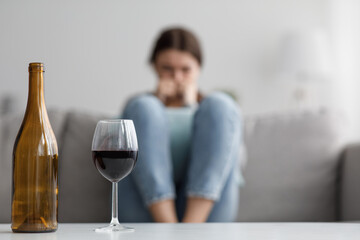 Sad caucasian millennial female sits on sofa suffering from stress and troubles, focus on bottle and glass of wine