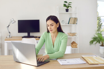 Portrait of young focused woman working with laptop and documents in open space office. Stylish young woman typing on laptop while sitting at desk on background of another workplace. Business concept.